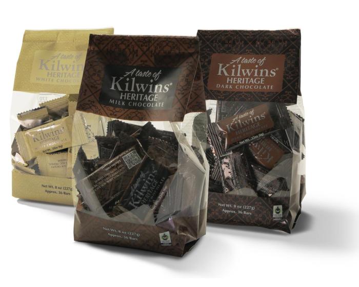 Three bags of Kilwins Heritage chocolates on a white background, including a gold bag of Spiced Chocolate and two dark brown bags of Milk and Dark Chocolate.